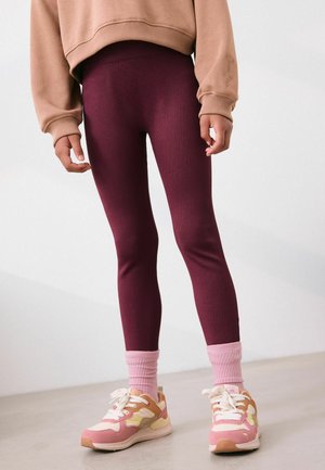 Person in ribbed burgunderfarbenen Leggings, rosa Socken und beigen und rosa Sneakers, die vor einem schlichten hellen Hintergrund steht.