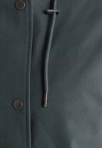 Tissu de veste Jack Wolfskin vert foncé avec cordon visible, fermoir métallique, logo embossé et boutons-pression le long de la couture.