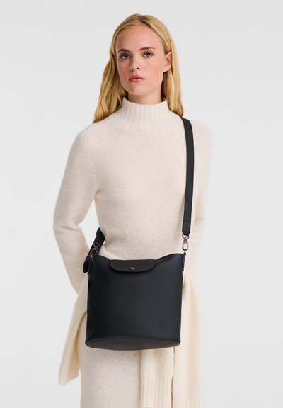 Frau mit langen blonden Haaren, die ein cremefarbenes Strickkleid trägt und eine schwarze Leder-Crossbody-Tasche mit einem Klappenverschluss trägt.