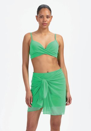 Femme debout, de face, portant un crop top vert à fines bretelles et un sarong transparent assorti noué à la taille.
