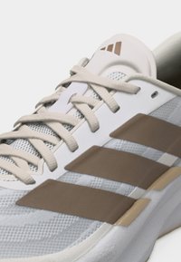 Άσπρο και μπεζ αθλητικό παπούτσι Adidas με μπεζ κορδόνια, τρεις μπεζ ρίγες στο πλάι και ανθεκτικό επάνω μέρος από διχτυωτό υλικό που επιτρέπει την αναπνοή.