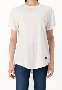 Biała bawełniana koszulka z krótkimi, podwiniętymi rękawami, okrągłym dekoltem i małym logo na piersi. Tył jest dłuższy.