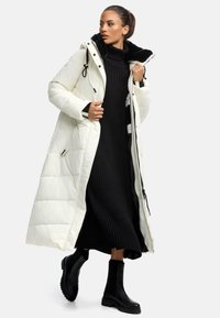 White puffer coat with a black faux fur hood, paired with a black ribbed dress and black chunky ankle boots. Zipper and drawstring details.