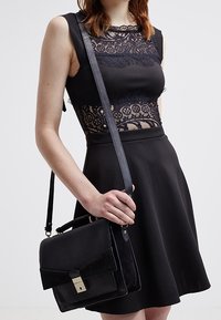 Czarna skórzana torba na ramię z zamszowym klapkiem, metalowym zapięciem i regulowanym paskiem; charakteryzuje się zarysowanym kształtem i minimalistycznym wzornictwem.