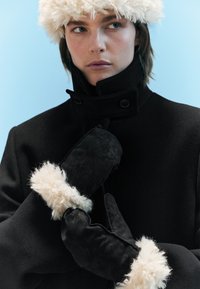 Black coat with a high collar, featuring button details. Black mittens with fluffy cream accents. Soft textures and tailored fit.