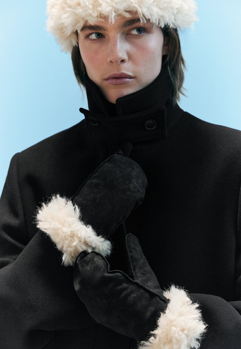 Black coat with a high collar, featuring button details. Black mittens with fluffy cream accents. Soft textures and tailored fit.