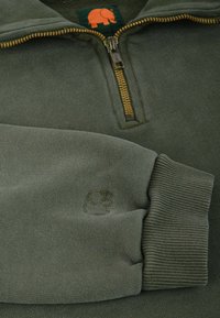 Sudadera verde oscura con media cremallera, puños acanalados y un logo bordado en la manga. Tejido suave con una textura lisa.
