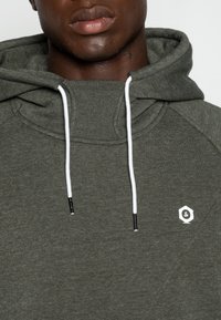 Felpa con cappuccio verde oliva in tessuto strutturato. Presenta un cordino con accenti bianchi e punte nere, e un piccolo logo bianco sul petto.