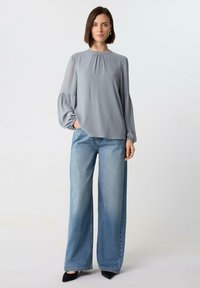 Helles graues Langarmbluse mit gerafftem Ausschnitt und voluminösen Ärmeln, kombiniert mit weiten blauen Denim-Jeans und schwarzen spitzen Absätzen.