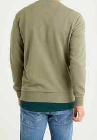 Sudadera verde oliva con puños y dobladillo acanalados, con diseño de cuello redondo. Usada sobre una camisa verde oscura y combinada con jeans azules.