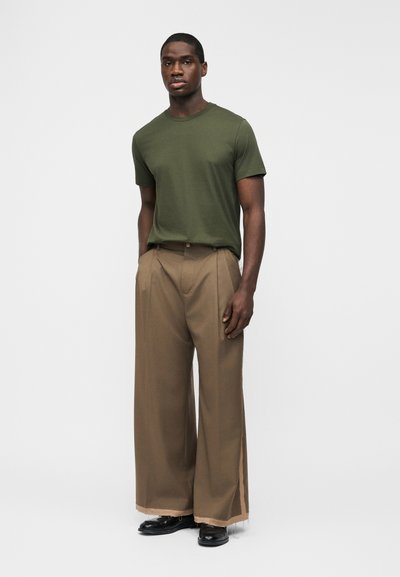 Άνδρας όρθιος, φορώντας ελιά κοντομάνικο πουκάμισο, μπεζ παντελόνι με φαρδιά μπατζάκια και ξεφτισμένα τελειώματα, και μαύρα παπούτσια, σε απλό ανοιχτό φόντο.