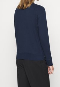 Marinblå sweatshirt, ribbade ärmslut och nederkant, rund halsringning, mjukt texturerat tyg, enkel design, bärs med svarta byxor, sedd bakifrån.