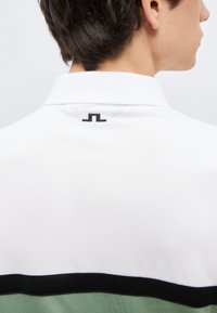 Άτομο φαίνεται από πίσω, φορώντας ένα λευκό και πράσινο πουκάμισο με γιακά, με ένα μικρό μαύρο λογότυπο στο πίσω μέρος του γιακά.