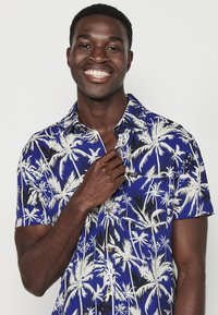 Camicia blu a maniche corte con bottoni, realizzata in tessuto leggero, con un motivo di foglie di palma bianche e una piccola tasca sul petto con un'etichetta.