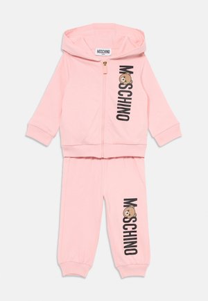 Ružičasti Moschino dječji trenirka sa kapuljačom na zatvarač i odgovarajućim tajicama. Crni logotip i grafika crtanog medvjedića na prednjoj strani. Mekani materijal, rebrasti završeci.