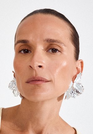 Nainen, jolla on taakse kammatut hiukset, suuri, kiiltäviä, hopeisia abstrakteja korvakoruja, neutraali meikki ja beige yläosa yksiväristä taustaa vasten.