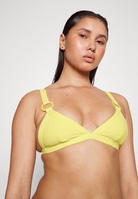 Top de bikini amarelo com riscas, design triangular, alças largas e detalhes em metal circular, apresentando uma textura suave e acabamento sem costuras.
