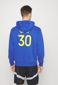 Felpa blu in cotone con cappuccio, con la scritta gialla "CURRY" e "30" sul retro, abbinata a pantaloni corti neri e dettagli grigi.