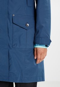 Blaue wasserdichte Jacke mit Brusttasche, Knopfdetail und Ärmelbündchen mit hellblauer Innenfutter. Glatte Textur, praktisches Design.