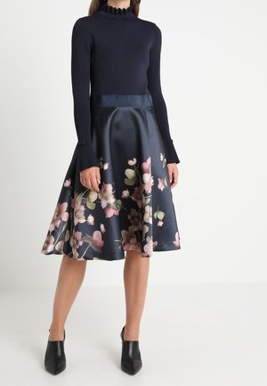 Vrouw draagt een marineblauwe top met lange mouwen en een satijnen rok tot op de knie met een roze bloemenprint, gecombineerd met zwarte enkellaarsjes met hak.