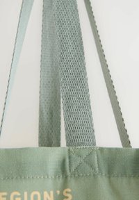 Borsa della spesa riutilizzabile in tessuto verde chiaro. Presenta manici intrecciati e cuciture cucite, con una superficie texture e testo stampato.