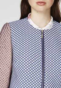Blue and white zip-up jacket with a diamond pattern on the body and a textured maroon mesh on the sleeves. Features a white collar.