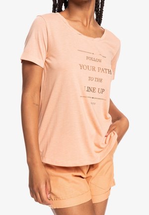 Personne portant un t-shirt pêche avec le texte "FOLLOW YOUR PATH TO THE LINE UP" et un short pêche assorti, main dans la poche, cheveux tressés visibles.