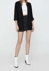 Woman wearing a black blazer, white crop top, black polka dot mini skirt, and white ankle boots standing against a plain background.