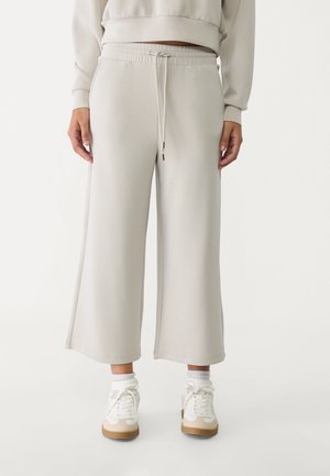 Osoba w jasnobeżowych, szerokich, krótkich spodniach z elastycznym pasem i sznurkiem, białych skarpetkach i białych trampkach.