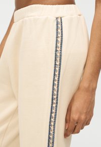 Κοντινό πλάνο μπεζ φόρμας με ελαστική μέση και κάθετη μπλε και μπεζ πλευρική ρίγα με κυματιστό μοτίβο, δείχνοντας ένα χέρι που ακουμπά δίπλα στο πόδι.