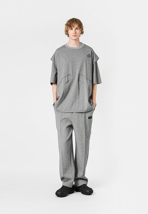 Camiseta gris de gran tamaño con diseño cuadrado y dos bolsillos frontales, combinada con pantalones grises que presentan un detalle de cadena lateral y puños elásticos.