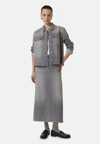 Veste en denim gris avec deux poches avant, assortie d'une jupe maxi en denim gris. Le modèle porte des mocassins noirs et des chaussettes grises.