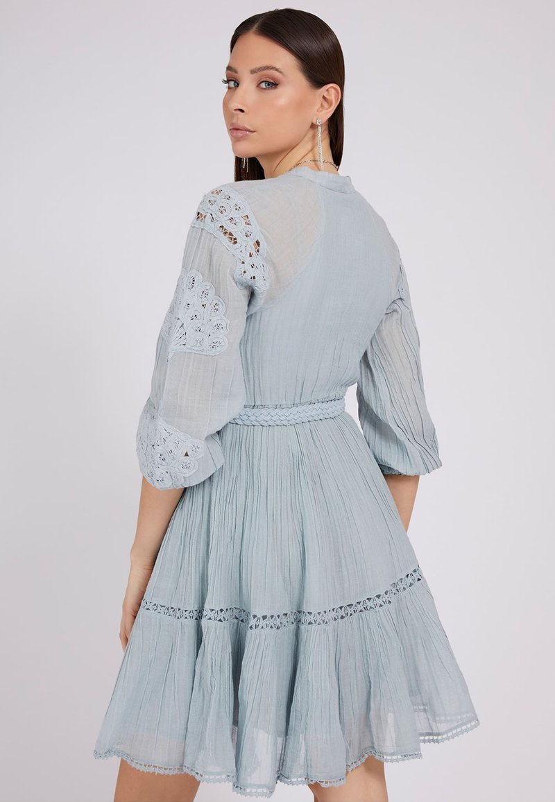 Vrouw met stijl bruin haar draagt een lichtblauwe jurk tot de knie met kantdetails, pofmouwen en een gevlochten tailleband, en kijkt over haar schouder.