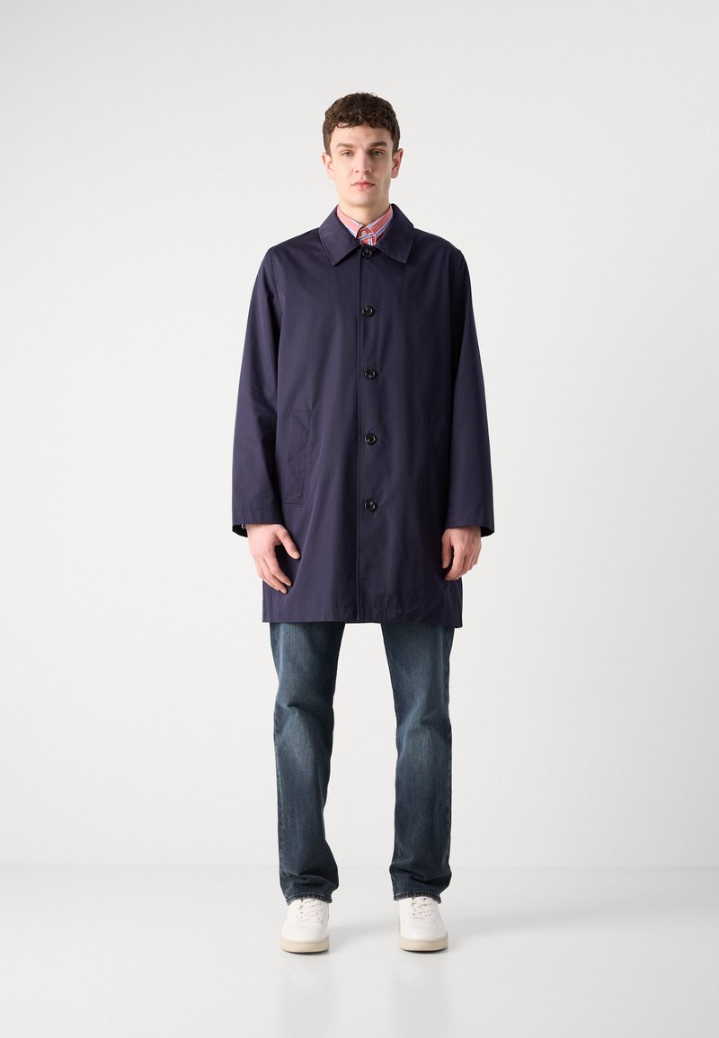 Granatowy wodoodporny płaszcz trench z prostym krojem, wyposażony w spiczasty kołnierz, sześć guzików z przodu i kieszenie po bokach. Noszony na dżinsy i buty.