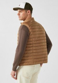 Gilet marrone imbottito con cuciture verticali, abbinato a una maglietta a maniche lunghe marrone. Un paio di pantaloni beige completa l'outfit.