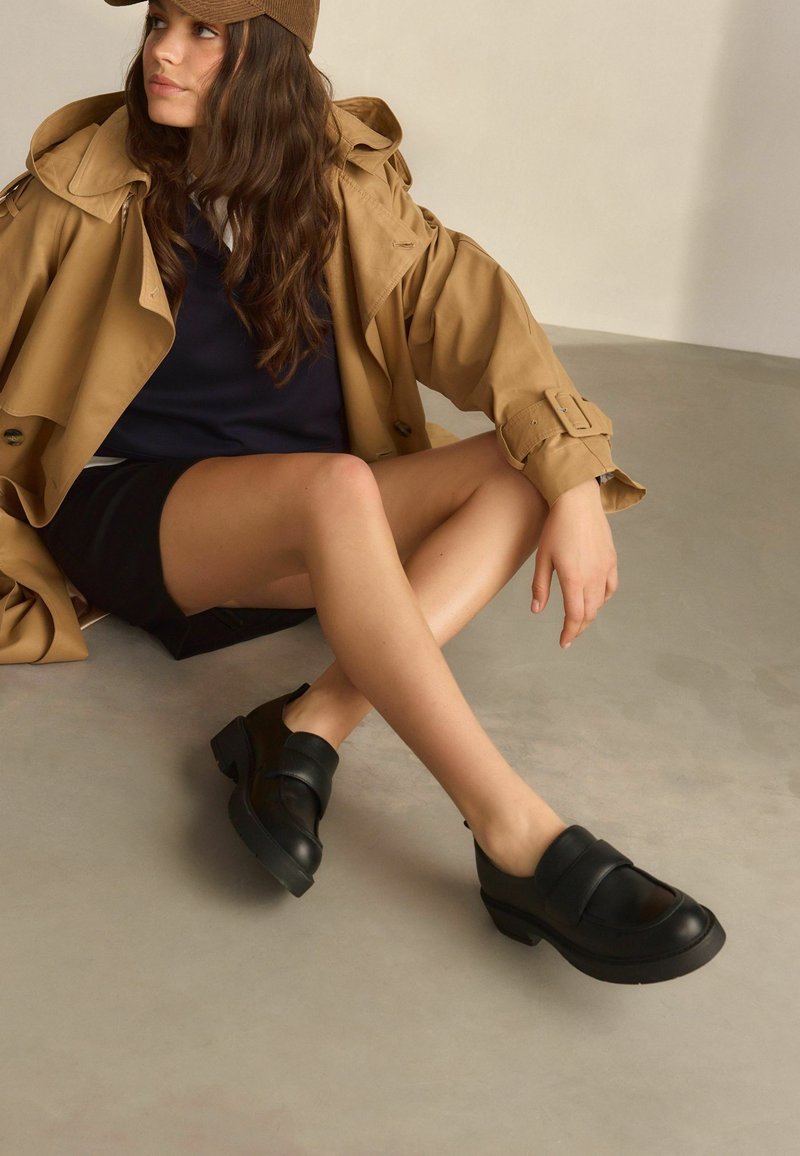 Black leather shoes with a rounded toe and strap detail, featuring a low chunky heel. Surface is smooth with a matte finish.