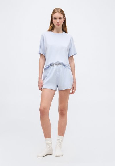 Νεαρή γυναίκα στέκεται φορώντας ανοιχτό μπλε μπλουζάκι με κοντά μανίκια και ταιριαστά σορτς με εμφανές λογότυπο στη μέση, λευκές κάλτσες μέχρι τον αστράγαλο, μπροστά σε απλό φόντο.