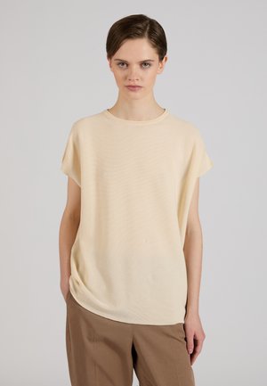 Jeune femme aux cheveux courts marron, portant un haut beige ample à manches courtes et un pantalon marron, debout devant un fond gris uni.