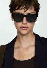 Occhiali da sole neri con un design geometrico e angolare. Lenti scure e montatura in plastica. Indossati con un top marrone e un blazer scuro. Stile minimalista.