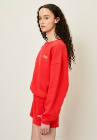 Röd sweatshirt och shorts set i mjukt tyg; oversized sweatshirt med långa ärmar, ribbad krage och liten logotyp på bröstet.