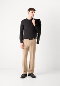 Chemise noire à manches longues, pantalon beige, chaussures en cuir noires. Le modèle tient une veste sur son épaule, se tenant contre un fond blanc.