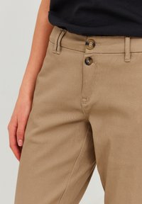 Pantalons khaki avec une texture lisse, dotés de deux boutons sombres à la taille et de poches latérales. Le tissu semble résistant.