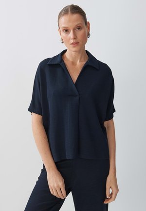 Blusa corta blu navy con colletto, vestibilità comfort, tessuto strutturato e dettaglio frontale a forma di V. Indossata con pantaloni coordinati.