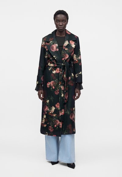 Femme portant un long manteau sombre à fleurs avec une ceinture, un jean large bleu clair et des chaussures noires pointues, debout devant un fond uni.