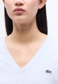 Jeune personne aux longs cheveux foncés portant un t-shirt blanc à col en V avec un petit logo de crocodile vert sur la poitrine.