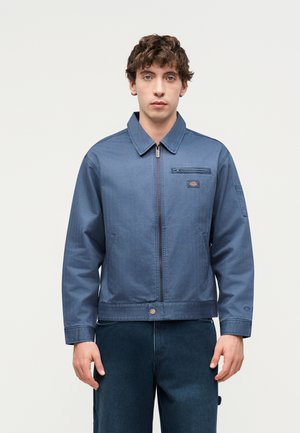 Junger Mann trägt eine blaue Arbeitsjacke mit Reißverschluss sowie Brust- und Seitentaschen und steht vor einem schlichten hellen Hintergrund.