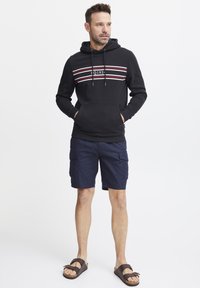 Felpa nera con strisce orizzontali multicolori, tasca a marsupio, abbinata a pantaloni cargo blu navy e sandali marroni.