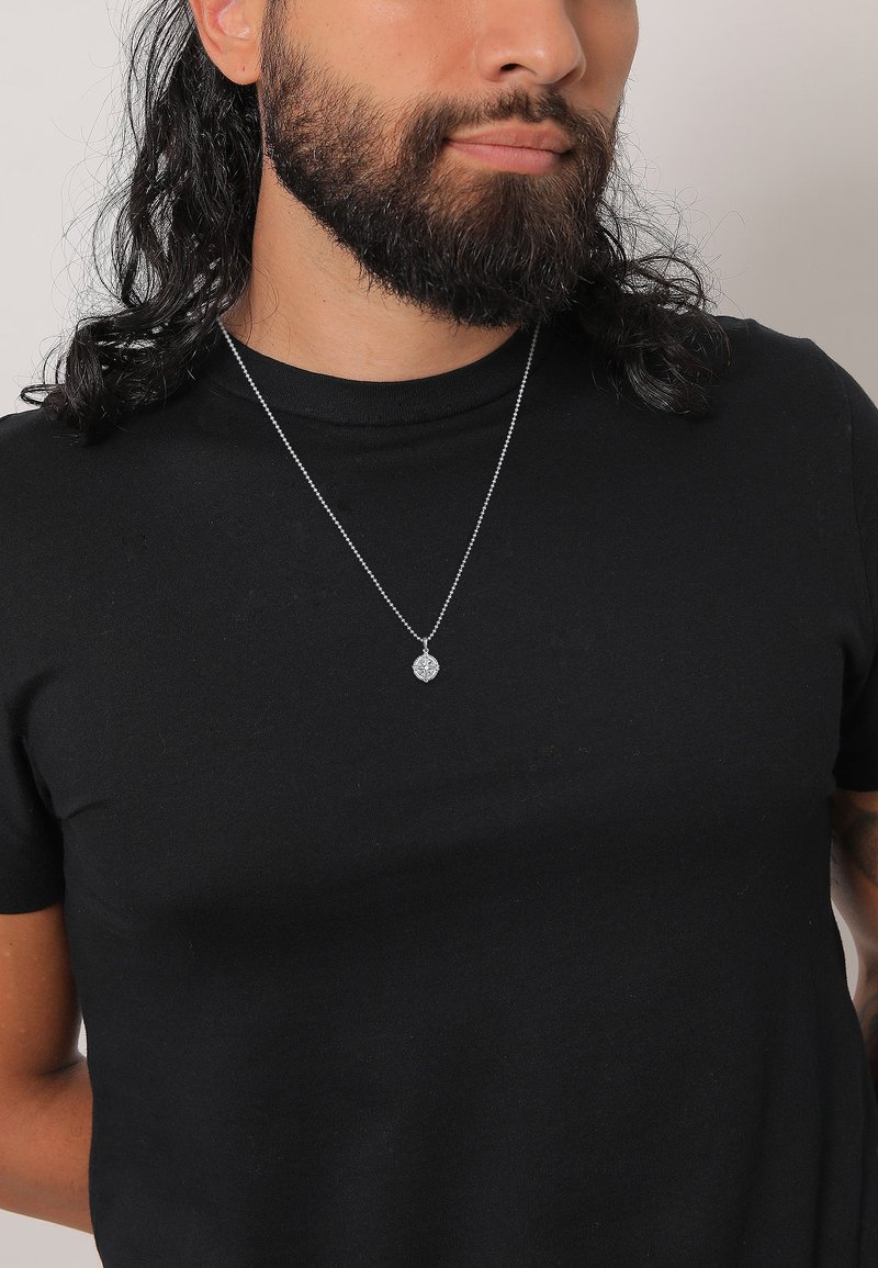 Silberner Anhänger an einer dünnen Kette, mit einem detaillierten Blumenmuster, getragen über einem schwarzen, kurzärmeligen T-Shirt.