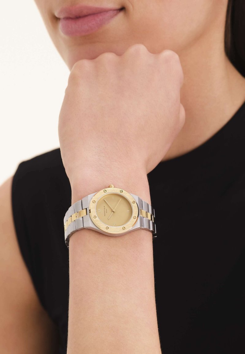 Femme portant une montre-bracelet en or et argent avec un cadran doré, reposant son poing près de son menton sur un fond clair.