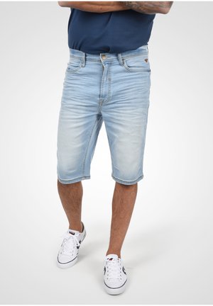 Hombre con pantalones cortos vaqueros de color azul claro, zapatillas blancas y una camisa azul oscuro con los brazos cruzados, de pie contra un fondo liso.